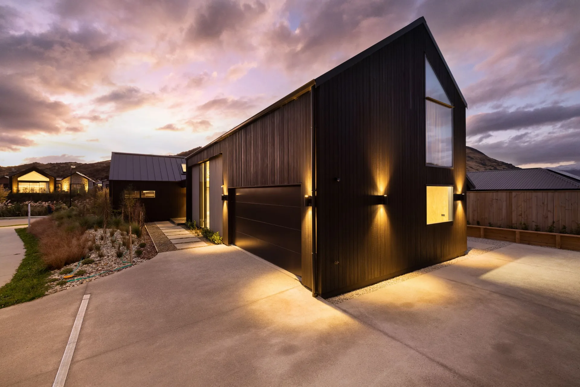 Finished black-clad Drysdale Road home with warm exterior lights beneath a dusk sky, Queenstown