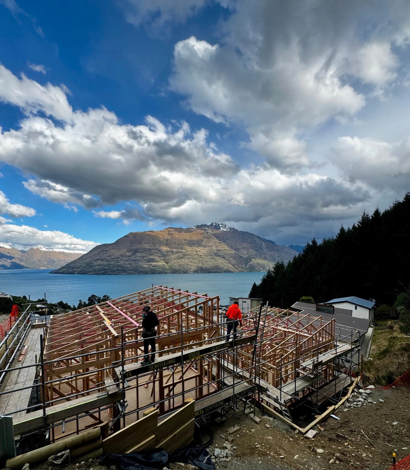 Te Tihi Villas roof framing on scaffolding with two builders, Lake Wakatipu and Cecil Peak beneath clouds