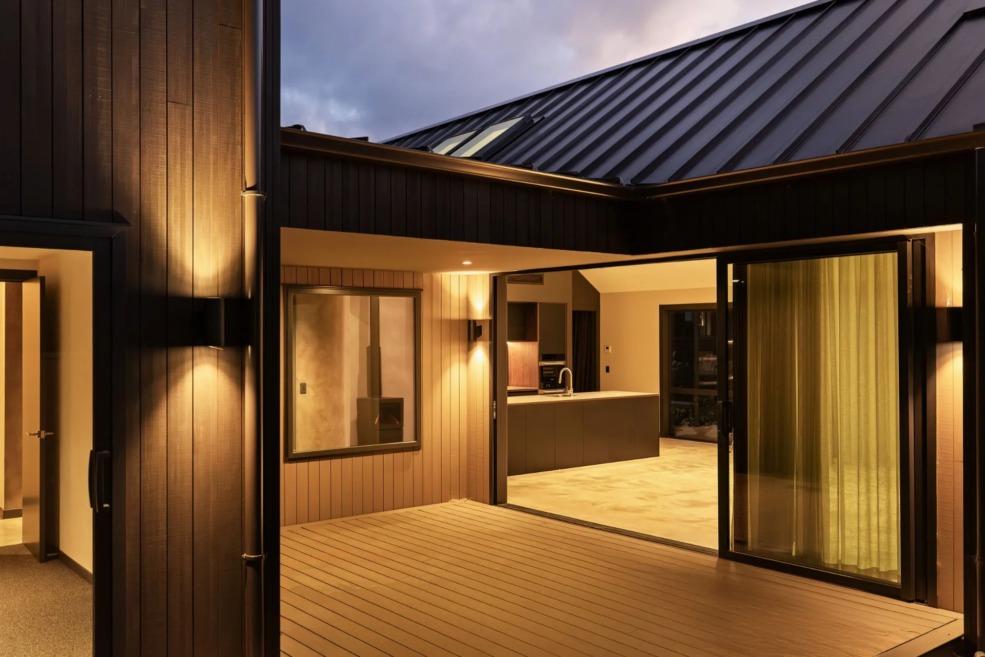 Open sliding doors from a timber deck into a warmly lit kitchen at dusk, Queenstown